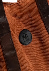 Brązowa torba typu tote z kordurowego materiału z ciemnobrązowymi skórzanymi uchwytami. Na boku znajduje się czarna okrągła naszywka z logo. Teksturowana powierzchnia z prążkowanym wzorem.