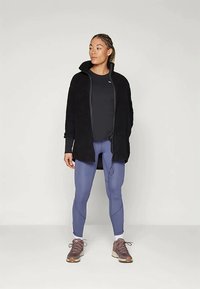 Γυναίκα στέκεται φορώντας μαύρο μπουφάν με φερμουάρ, μαύρη μακρυμάνικη μπλούζα, μπλε κολάν, λευκές κάλτσες και καφέ αθλητικά παπούτσια σε απλό φόντο.