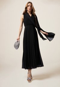 Zwarte, vloerlange jurk met geplisseerde rok en aansluitende taillering. Heeft een brede sjaal die over één schouder hangt. Zilveren gespsandalen en een kralenclutch.