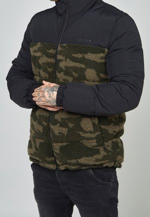 Homme portant une veste polaire camouflage verte avec des manches noires rembourrées, les mains jointes devant lui, montrant des doigts tatoués.