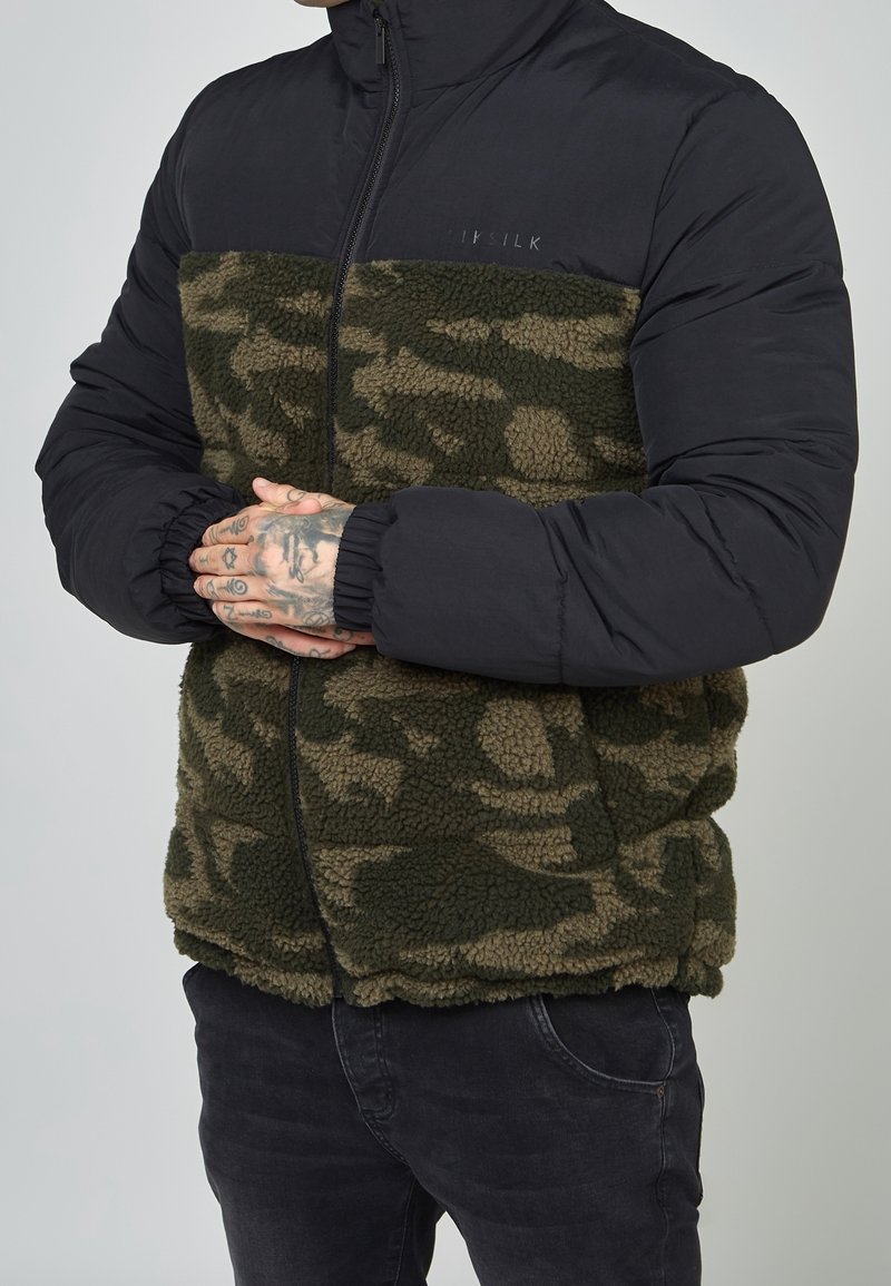 Homme portant une veste polaire camouflage verte avec des manches noires rembourrées, les mains jointes devant lui, montrant des doigts tatoués.