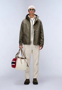 Olivgrüne wasserdichte Jacke, cremefarbener Pullover, beige Cargohose, dunkelbraune Stiefel, trägt eine creme-rote Reisetasche mit Trägern.