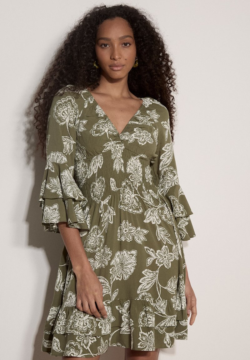 Femme aux longs cheveux bouclés portant une robe fleurie vert olive avec des manches à volants trois quarts et un col en V sur un fond uni.