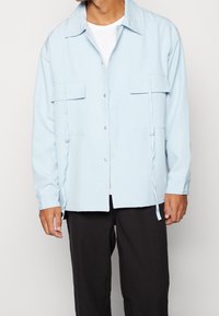 Ljust blå skjorta med krage, två bröstfickor, sidodetaljer med snöre och långa ärmar, tillverkad av ett slätt tyg.