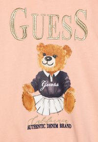 Rosa stoff med en bjørn i en svart polo med "Guess" brodert og et hvitt skjørt; gullbokstavene lyder "CALIFORNIA AUTHENTIC DENIM BRAND."
