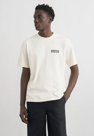 Hombre de piel oscura vistiendo una camiseta blanca de manga corta con el logo "Dickies" en el pecho y pantalones negros, de pie frente a un fondo claro y liso.