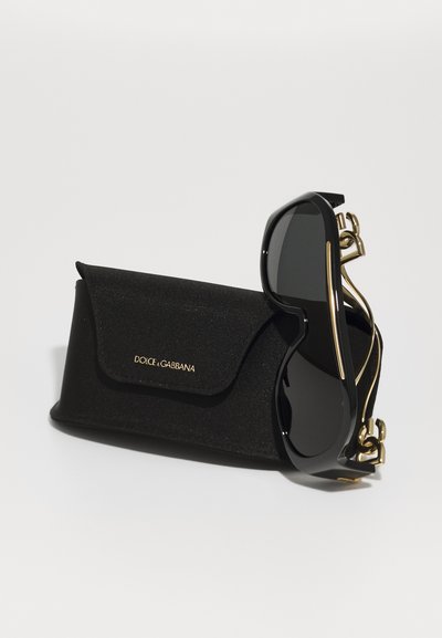 Schwarze Sonnenbrille mit dunklen Gläsern, goldenen Akzenten und einer einzigartigen Form, neben einem strukturierten schwarzen Etui mit Logo.