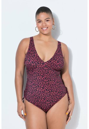 Lächelnde Frau mit kurzen Haaren trägt einen rot-schwarz leopardenmuster Badeanzug, goldene Creolen und leuchtend orangefarbene Nagellack, steht vor einer weißen Wand.