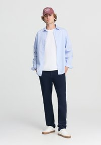 Camicia button-up azzurro chiaro, t-shirt bianca, pantaloni blu navy, accenti marroni. Cappellino bordeaux con il testo "New Jersey East Coast", sneakers bianche.