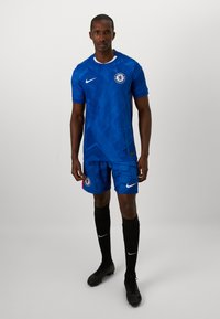 Domowy zestaw piłkarski Chelsea FC w kolorze niebieskim, z geometrycznym wzorem. Koszulka ma biały kołnierzyk i logo; szorty ozdobione są emblematem klubu. Czarne skarpety i korki.