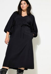 Černé šaty do kolen s výstřihem do V, nafouknutými rukávy a stahovacími šňůrkami v pase. Mají hladkou texturu a jednoduchý, minimalistický design.