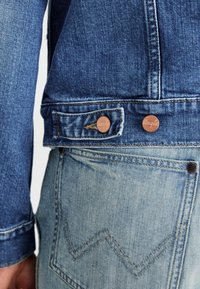 Jeansowa kurtka w ciemnoniebieskim kolorze, z mosiężnymi guzikami, oraz jasnoniebieskie dżinsy z wystającym wzorem w zygzak.