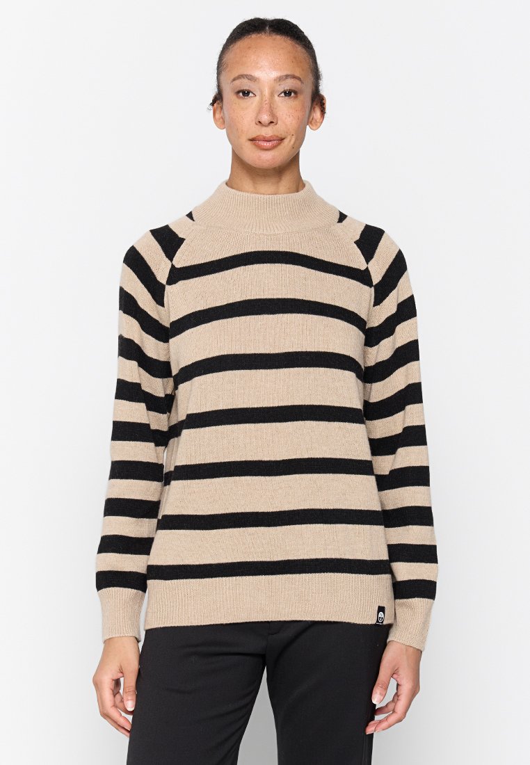 Donna con i capelli legati, che indossa un maglione a righe orizzontali beige e nero e pantaloni neri, in piedi davanti a uno sfondo bianco semplice.