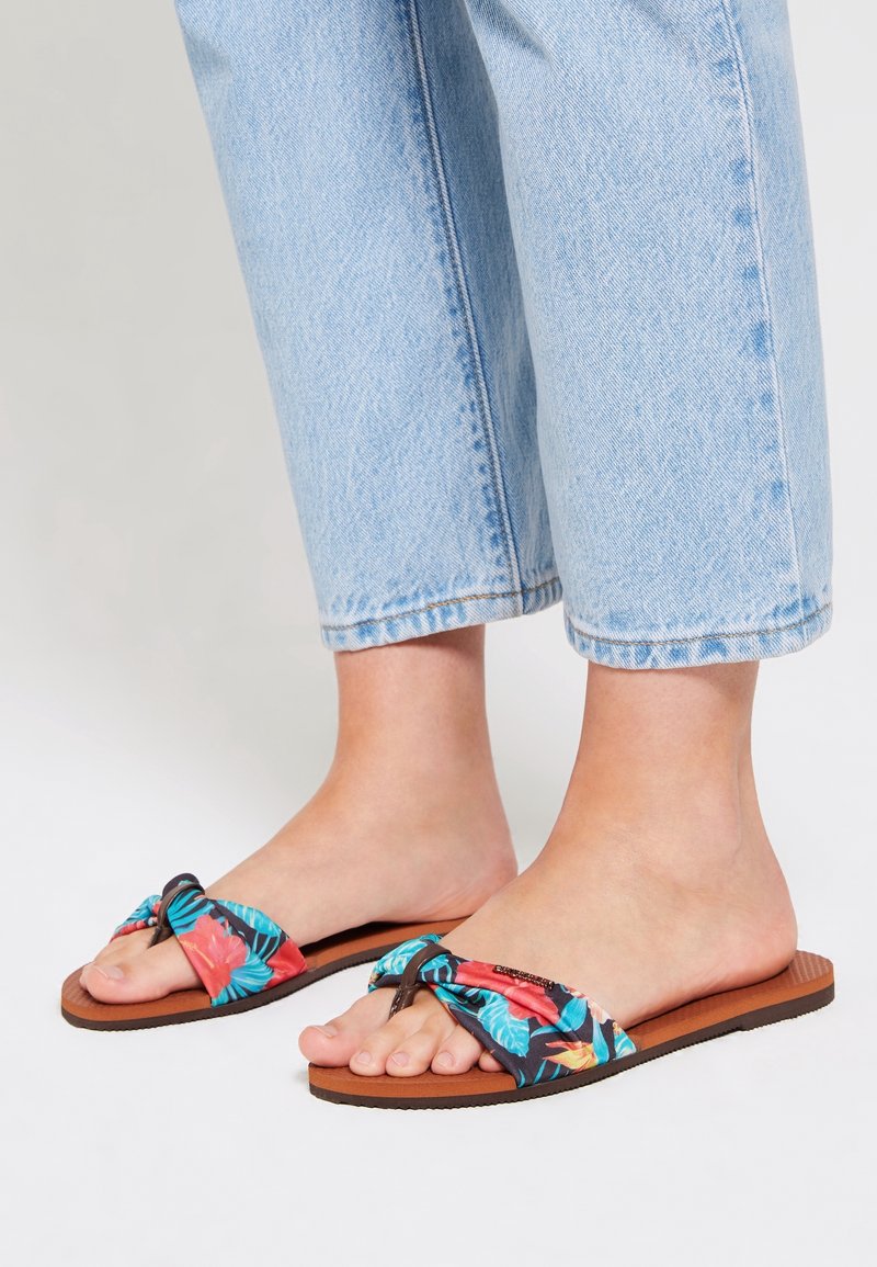 Pieds portant un pantalon en denim bleu clair coupé court et des sandales plates marron avec des sangles en tissu floral coloré.
