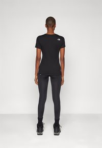 Camisa preta de manga curta com um pequeno logotipo branco na parte superior das costas, combinada com leggings pretas e sapatilhas desportivas cinzas, vista pela parte de trás.