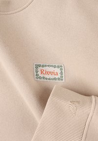 Beiger Sweatshirt mit geripptem Kragen und Bündchen, mit einem rechteckigen Etikett, auf dem "Rivvia" in Orange steht, umgeben von grünen floralen Akzenten.