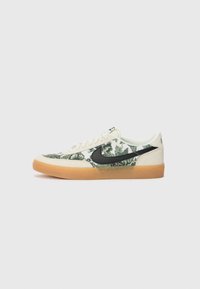 Sneaker basse con bordi in suede beige, tessuto floreale verde, logo Nike nero, lacci bianchi e suola in gomma marrone su sfondo bianco.