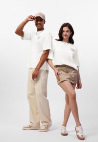 To modeller har på seg uformelle antrekk: en hvit oversized t-skjorte med grafikk, beige cargo-shortser og matchende sneakers. Den mannlige modellen har på seg en caps.