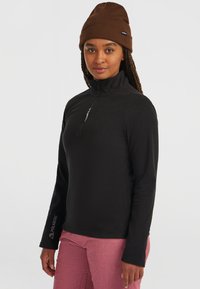 Zwarte fleecetop met een halve ritssluiting, met een Polartec-logo op de mouw. Gemaakt met roze broek en een bruine gebreide muts.