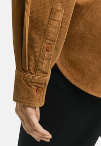 Brown corduroy shirt with textured fabric, featuring buttoned cuffs and orange buttons. Visible stitching detail near the cuff.