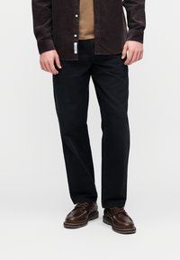 Temno modre hlače s krojem s pravo nogavico, kombinirane s svetlo rjavo majico in črno kordurovo srajco. Obleko zaključujejo rjavi čevlji z vezalkami.