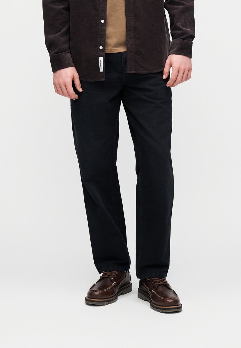 Pantaloni blu scuro a gamba dritta, abbinati a una T-shirt marrone chiaro e una camicia di velluto a coste nera. Scarpe marroni con lacci completano il look.