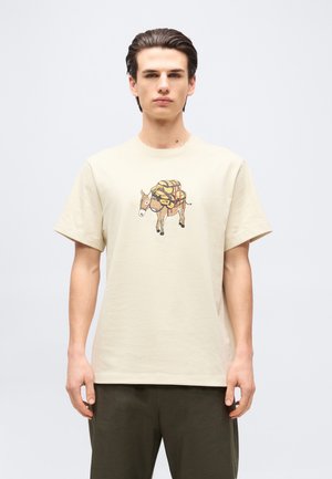 Joven con camiseta beige con una ilustración de un burro marrón cargando paquetes amarillos, posando frente a un fondo liso.