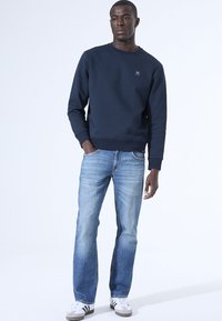 Marineblauwe sweatshirt met een gestructureerd patroon, ronde hals, ribgebreide manchetten; gedragen met lichtblauwe jeans en witte sneakers met zwarte strepen.