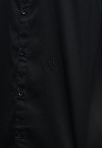 Μαύρο πουκάμισο με κουμπιά και διακριτικό κεντητό λογότυπο "A|X" στο στήθος, με τέσσερα εμφανή μαύρα κουμπιά μπροστά.