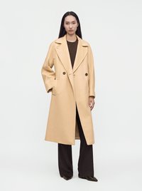 Femme aux longs cheveux noirs portant un manteau beige à double boutonnage sur un pantalon noir et des chaussures noires pointues, debout devant un fond blanc uni.