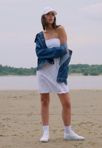 Hvit kjole med blonder, kombinert med en stor denimjakke, hvite sokker og hvite joggesko. Plassert på en sandstrand med grønne områder i bakgrunnen.