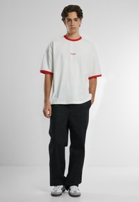 T-shirt bianca a maniche corte con polsini rossi e logo ricamato, abbinata a pantaloni neri a gamba larga e sneakers grigie.