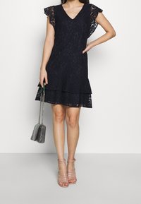 Marinblå spetsklänning med V-ringning och korta volangärmar, med en flikig kjol. Modellen håller en grå handväska och har på sig genomskinliga klackar.