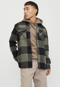 Camisa de cuadros verde y negra con botones en el frente, dos bolsillos en el pecho y mangas largas, superpuesta sobre una sudadera beige con capucha.