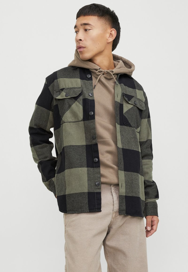 Camisa de cuadros verde y negra con botones en el frente, dos bolsillos en el pecho y mangas largas, superpuesta sobre una sudadera beige con capucha.