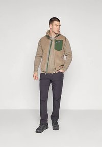 Κιτρινόθριχης fleece ζακέτας με πράσινη τσέπη στο στήθος, κλείσιμο με φερμουάρ και κουκούλα. Συνδυασμένη με μαύρο παντελόνι και μαύρα παπούτσια. Απλός, λειτουργικός σχεδιασμός.