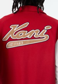 Röd fleecejacka med en stor broderad "Kani"-logotyp i beige och vitt, med texten "est. 1989" nedanför. Slät textur.