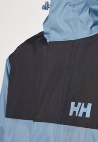 Waterproof jacket with a colorblocked design in light blue and black. Features a hood and the "HH" logo in light blue on the chest.