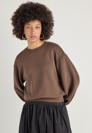 Felpa marrone oversize in un tessuto morbido, con scollatura rotonda e spalle cadenti, abbinata a una gonna nera arricciata.