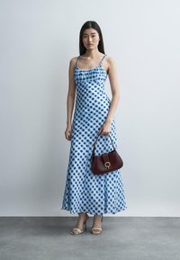 Ženska, ki nosi dolgo modro-belo karo obleko, drži temno rjavo torbico in nosi bež sandale z visokimi petami na sivi talni površini.