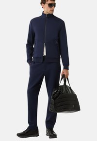 Granatowy dres na zamek błyskawiczny z teksturowanej tkaniny, z prążkowanymi mankietami i pasem. Czarna kurtka puchowa trzymana w jednej ręce; eleganckie czarne sneakersy.