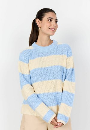 Woman with dark hair in a ponytail wearing a blue and cream striped knit sweater, beige pants, and gold earrings, smiling and looking right.