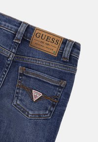 Jeans de mezclilla en azul oscuro con una etiqueta de marca de cuero, que presentan un logotipo triangular en el bolsillo trasero y detalles de costura en contraste.