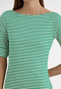 Žena oblečená v zeleno-bielom tričku s horizontálnymi pásmi, krátkymi rukávmi a širokým výstrihom loďkového tvaru na bielom pozadí.