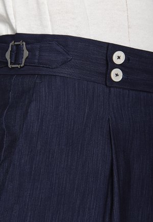 Pantaloni a righe sottili blu scuro con doppi bottoni bianchi e dettaglio con fibbia laterale in vita, indossati con una maglia beige chiaro infilata dentro.