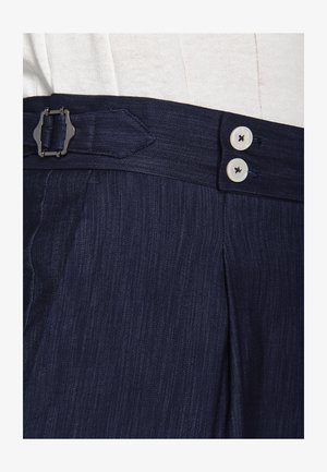 Pantalones de rayas azul oscuro con doble botón blanco y detalle de hebilla lateral en la cintura, combinados con una blusa beige clara metida por dentro.