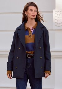 Dubbelgeknoopte marineblauwe jas met gouden knopen, gecombineerd met een meerkleurige gestreepte trui in bruin, blauw en roze. Donkere spijkerbroek maakt de outfit af.