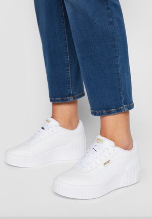 Osoba nosząca białe sneakersy Puma na platformie w połączeniu z przyciętymi niebieskimi dżinsami na jednolitym tle.