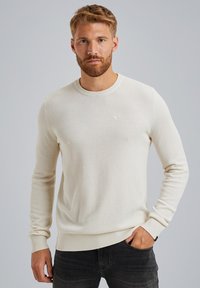Crèmekleurige gebreide trui met een gestructureerd patroon, ronde halslijn en geribde manchetten. G draagt over donkere jeans, weergegeven tegen een neutrale achtergrond.