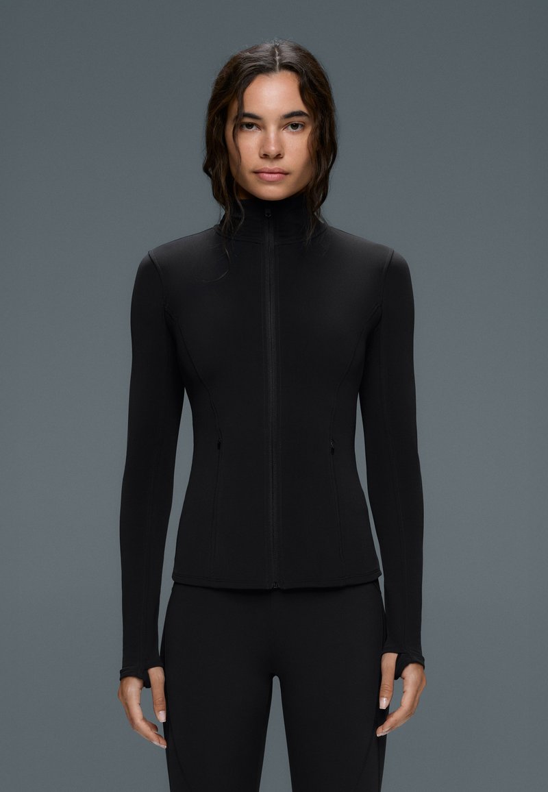 Femme aux cheveux bruns ondulés portant une veste noire ajustée avec fermeture éclair et un pantalon noir assorti, debout devant un fond gris.